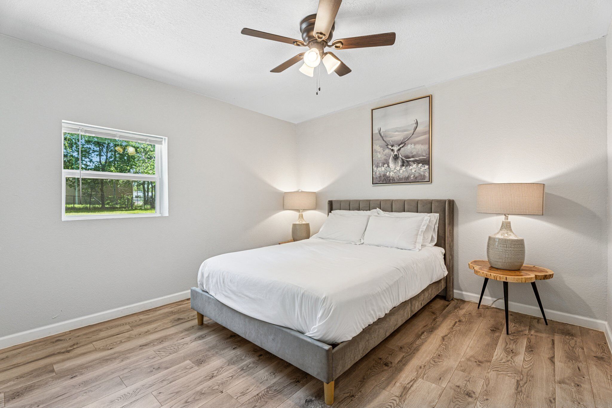 A bedroom with a bed, two bedside lamps, a ceiling fan, a window with a view, and a framed picture on the wall.