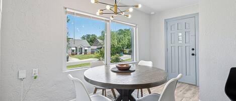 A dining area with a round table, four white chairs, a modern chandelier, and a view of the outside through large windows.