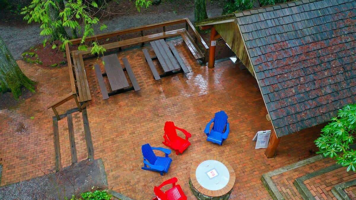 A covered outdoor seating area with wooden benches and chairs, a round table, and a brick floor.