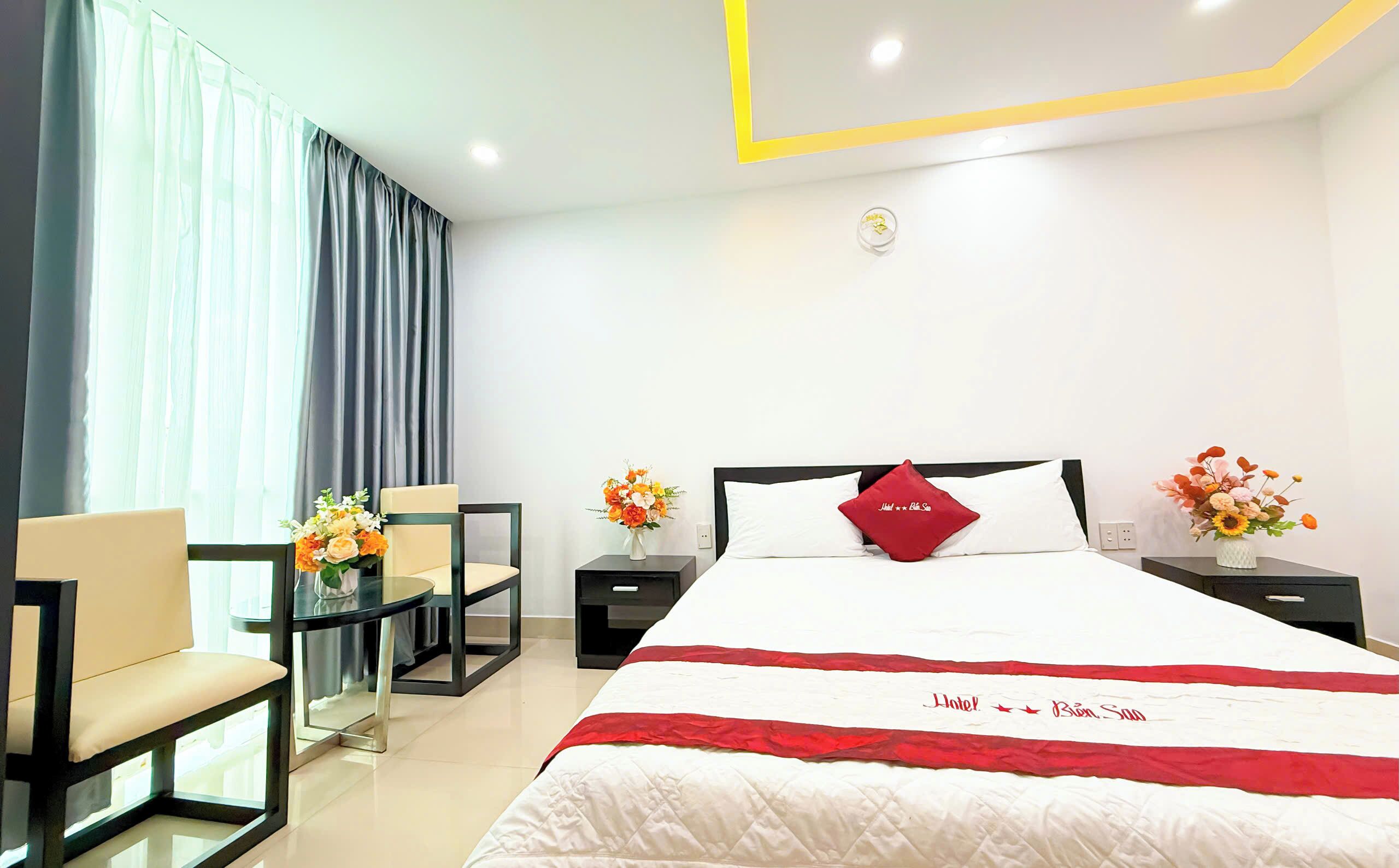 A hotel room with a bed, a desk, and chairs. There are flower arrangements on the nightstands.