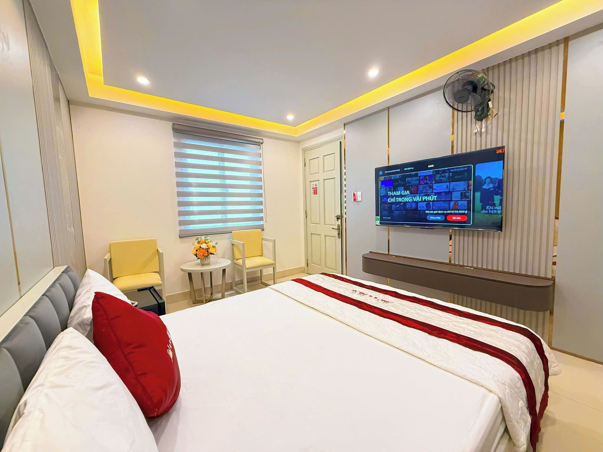 A hotel room with a large bed, a TV mounted on the wall, a small table with chairs, and a window with blinds.