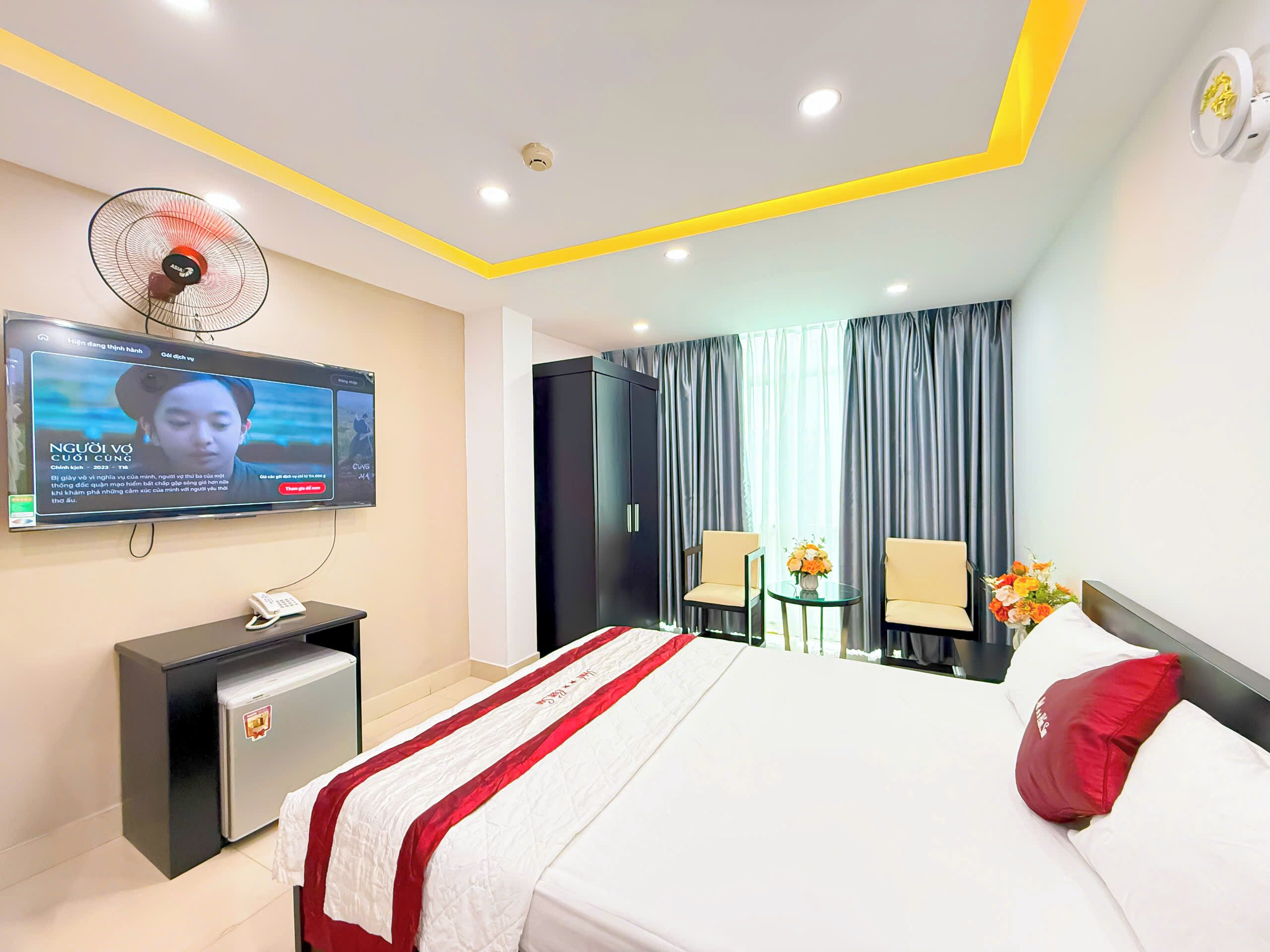 Une chambre d’hôtel avec un lit, un téléviseur fixé au mur, un petit bureau, un ventilateur et une armoire.