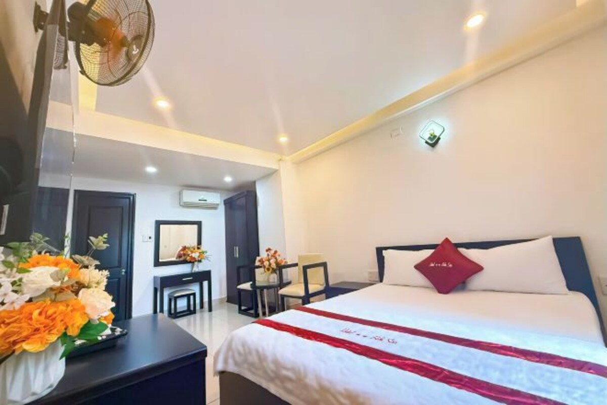 Quarto de hotel com cama, escrivaninha, cadeira, mesinha, espelho e ventilador.