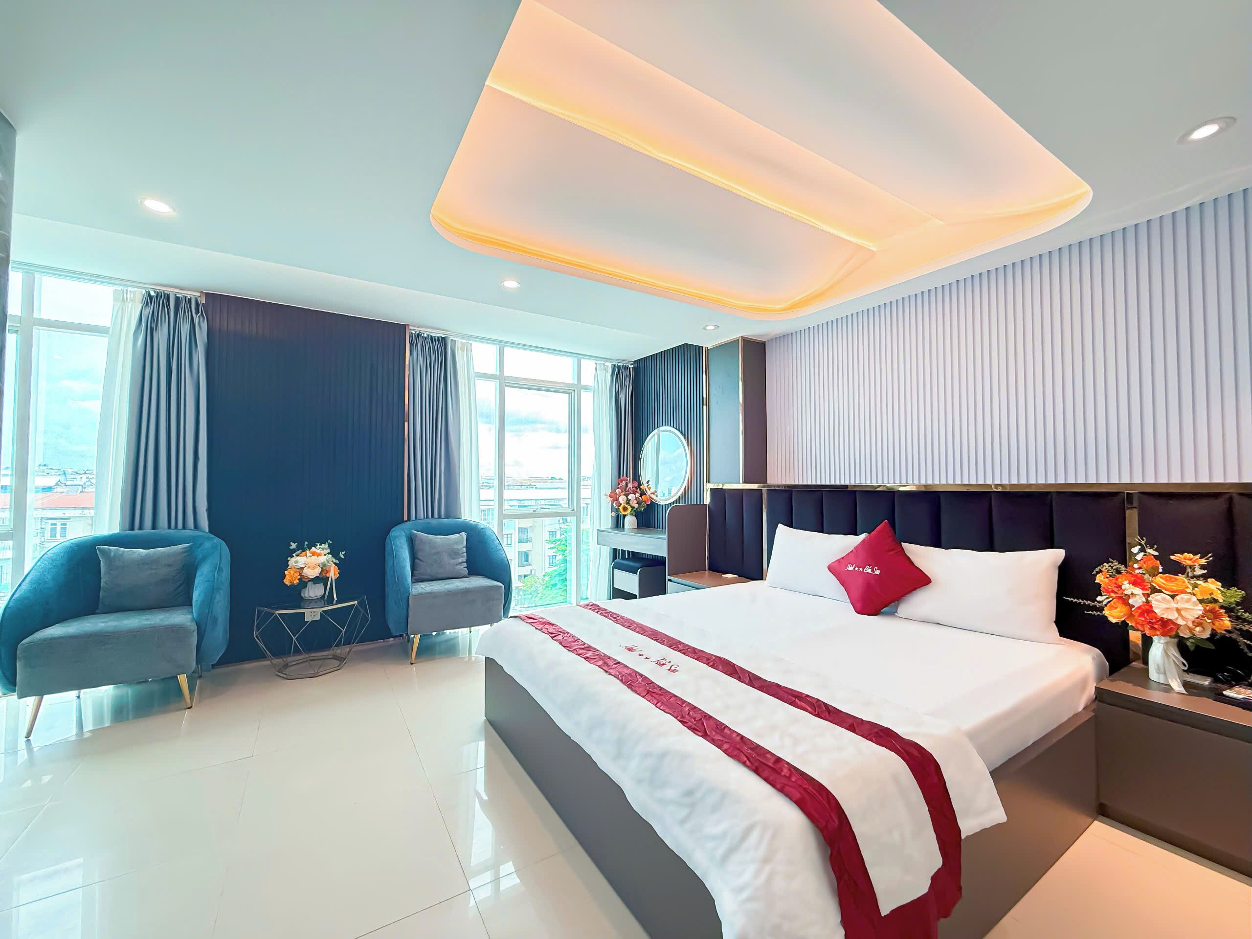 A modern hotel room with a large bed, a seating area with two armchairs, a desk, and a view of the city through large windows.