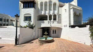 A white building with a fountain in front, featuring arched windows and a tiled entrance.