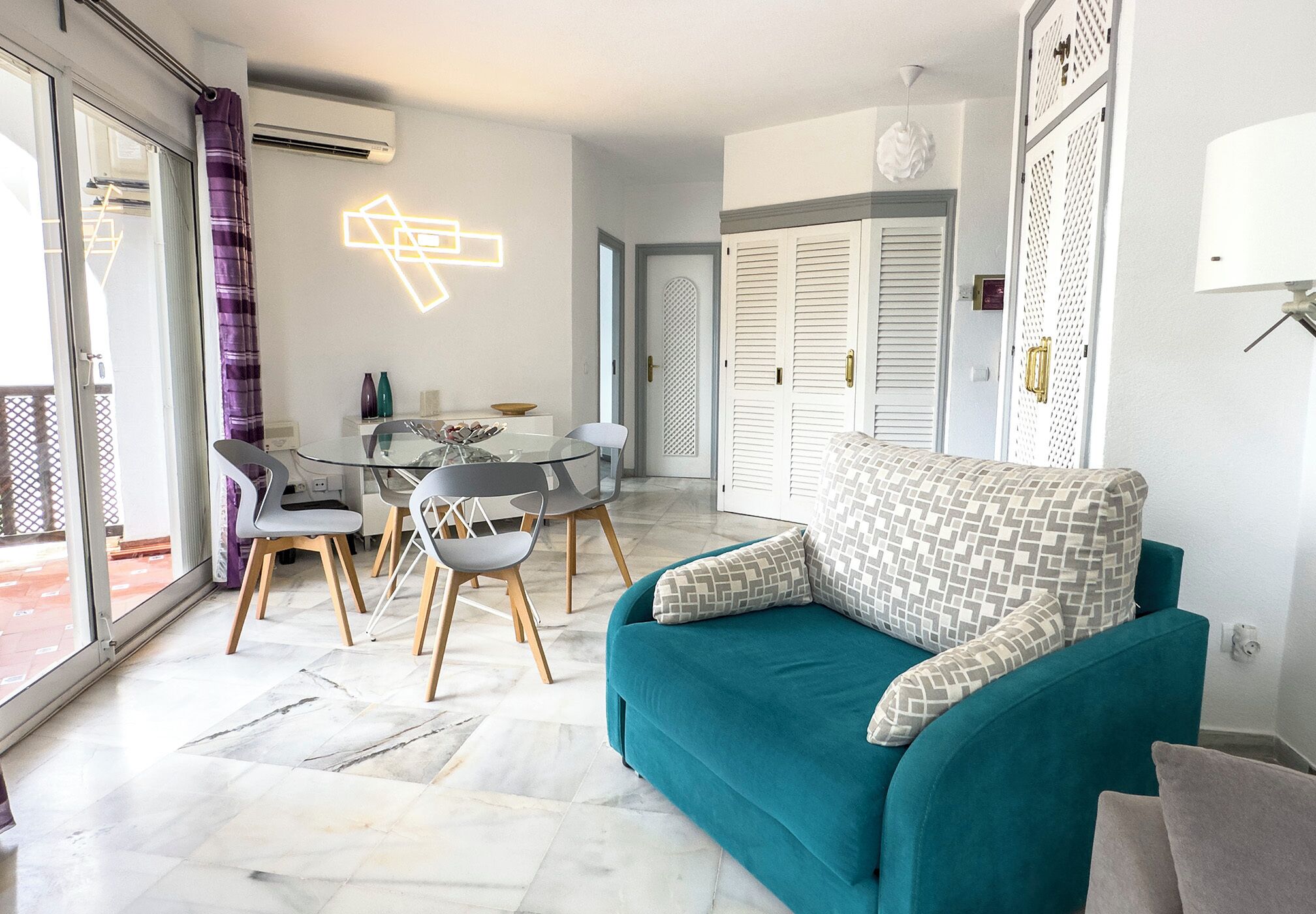 A modern living room with a teal sofa, a glass dining table with chairs, and a patterned throw pillow.