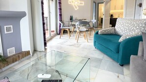 A modern living room with a teal sofa, a glass coffee table, and a dining area with a table and chairs.