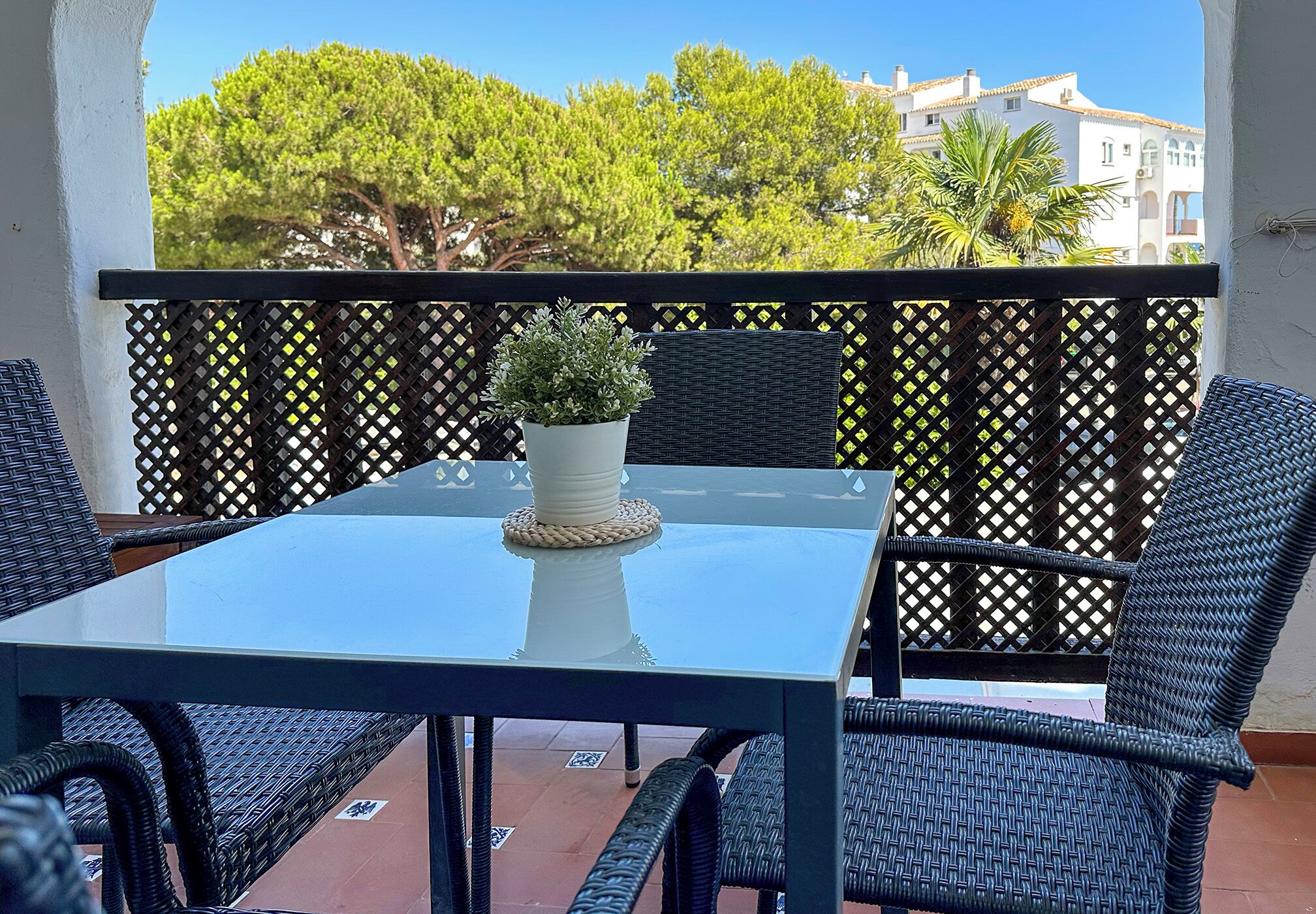 A balcony with a glass table and wicker chairs, a potted plant, and a view of trees and buildings.