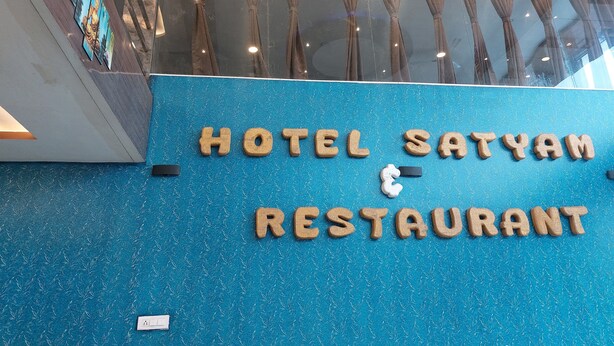 Skilt for Hotel Satyam Restaurant med træbogstaver på en blå, struktureret væg.