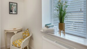 Une pièce avec une chaise en osier blanche, une petite table blanche, une plante en pot et une fenêtre avec des stores.