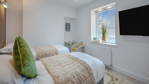 Une chambre avec deux lits, un radiateur, une fenêtre avec des stores, une petite table et un téléviseur fixé au mur.