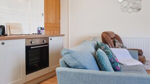 A modern kitchen with white cabinets, a built-in oven, and a wooden door. A blue sofa with decorative pillows is positioned in front of a radiator. - Pheasant Pens 1 - UK50187 (Llandenny, near Usk)