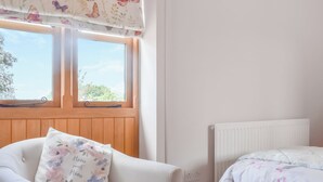 A bedroom with a bed, a chair with a floral pillow, a window with a butterfly curtain, and a white radiator. - Pheasant Pens 1 - UK50187 (Llandenny, near Usk)