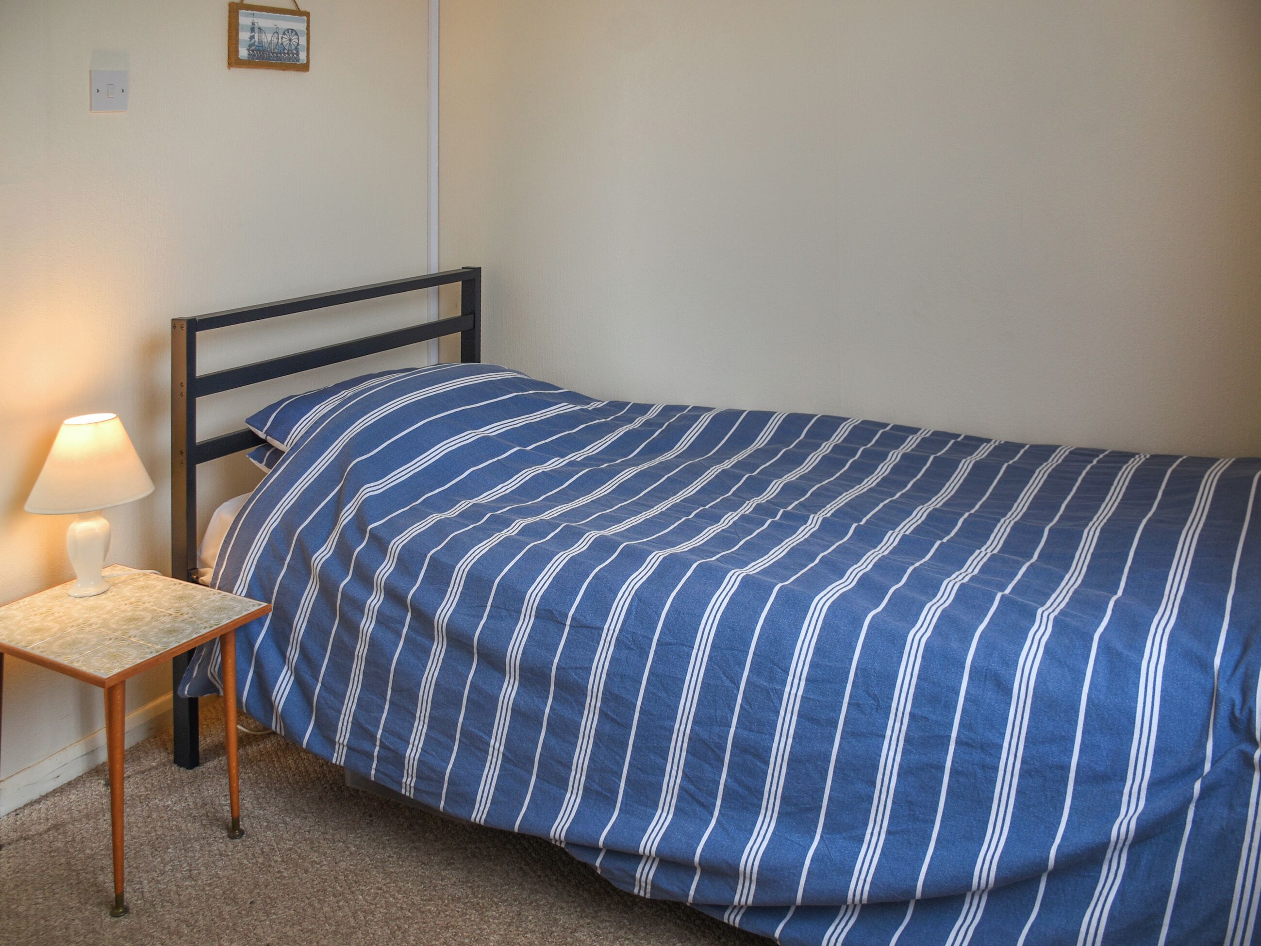 Een enkel bed met een blauw-witte streepdeken, een nachtkastje met een lamp, en een lijst met een foto aan de muur.