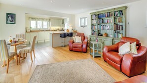 A spacious living area with a kitchen, a red leather sofa, a dining table with striped chairs, and a bookshelf filled with books.