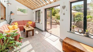Ein sonnendurchflutetes Zimmer mit Holzbalkendecke, einer Couch mit Kissen, einem Tisch und Blick auf einen Garten durch große Fenster.