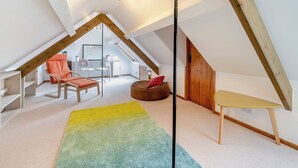 Ein Schlafzimmer im Dachgeschoss mit schrägen Wänden, Holzbalken, einem bunten Teppich, einem Bett, einem Schreibtisch und einem Stuhl.