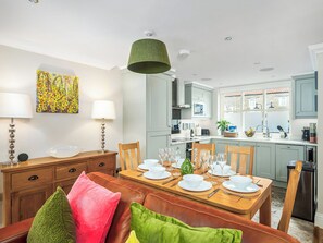 Uma sala de jantar com uma mesa de madeira posta para quatro pessoas, uma cozinha ao fundo e uma luminária pendente verde sobre a mesa.