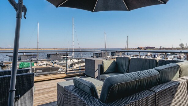 A deck with outdoor seating and a large umbrella, overlooking a marina with boats and a distant windmill. - Modern apartment with free view over the water (Uitgeest)