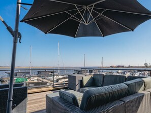 A deck with outdoor seating and a large umbrella, overlooking a marina with boats and a distant windmill. - Modern apartment with free view over the water (Uitgeest)