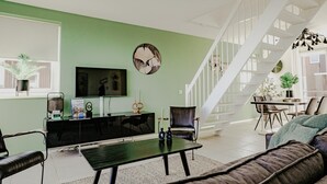 A modern living room with a green wall, a flat-screen TV, a black entertainment unit, a staircase, and a dining area with a table and chairs. - Modern apartment with free view over the water (Uitgeest)