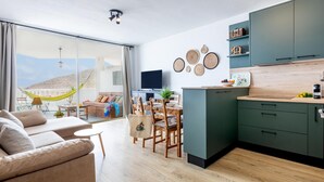 A modern living space with a sofa, dining table, chairs, a kitchen with green cabinets, and a large window overlooking a scenic view. - Modern 2-bedroom apartment with pool (Costa Adeje)
