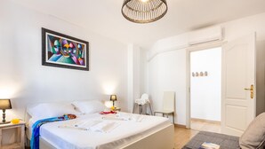 A neatly made bed with pillows and a blanket, a bedside table with a lamp, a chair, a small table with a book and glasses, and a door with a handle. - Modern 2-bedroom apartment with pool (Costa Adeje)
