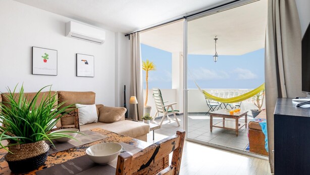 A modern living room with a sofa, dining table, and chairs, a balcony with a hammock, and a view of the sky. - Modern 2-bedroom apartment with pool (Costa Adeje)