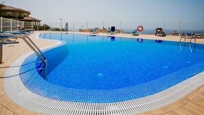 A clear blue pool with lounge chairs and a lifebuoy, surrounded by a tiled deck and a building in the background. - Modern 2-bedroom apartment with pool (Costa Adeje)