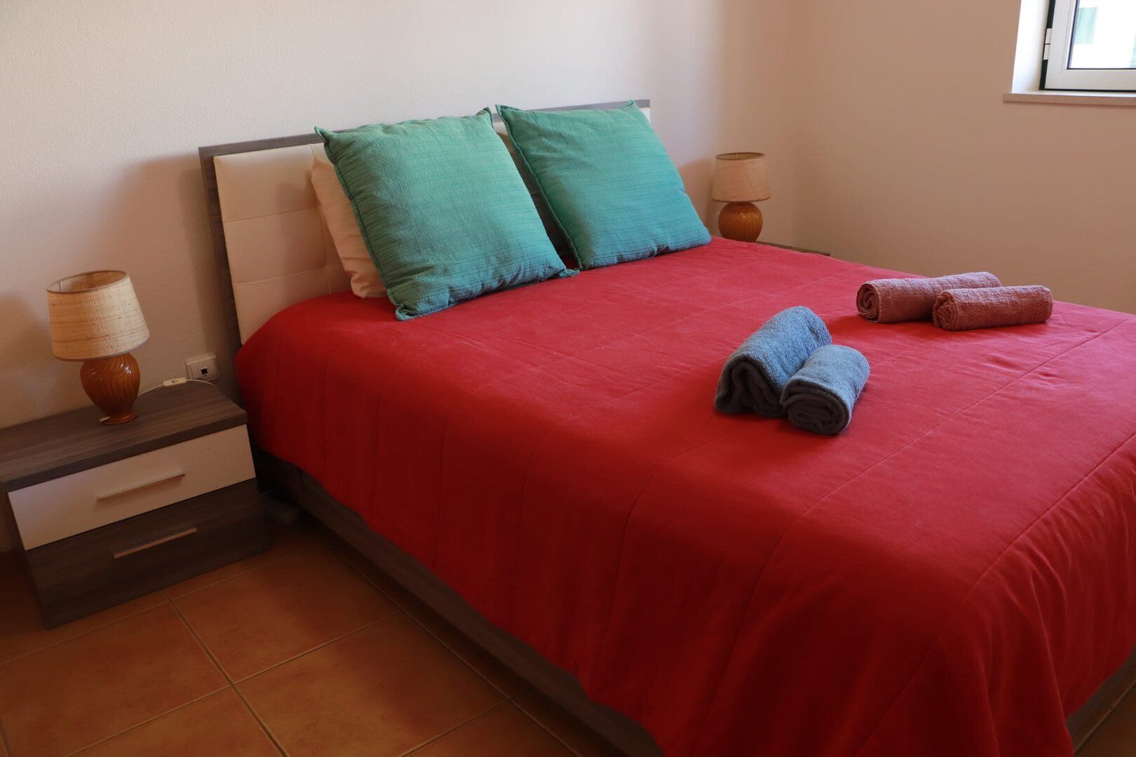 Un lit bien fait, avec une couverture rouge, deux coussins turquoise et deux serviettes enroulées.