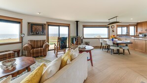 Ein geräumiger Wohnbereich mit großem Fenster mit Blick auf den Strand, ein Essbereich mit Küche im Hintergrund und eine gemütliche Sitzecke mit gemusterten Kissen.