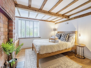 A bedroom with a wooden bed, a nightstand, a lamp, a window with blinds, and a plant. - 4 bedroom accommodation in Welshpool (Welshpool)