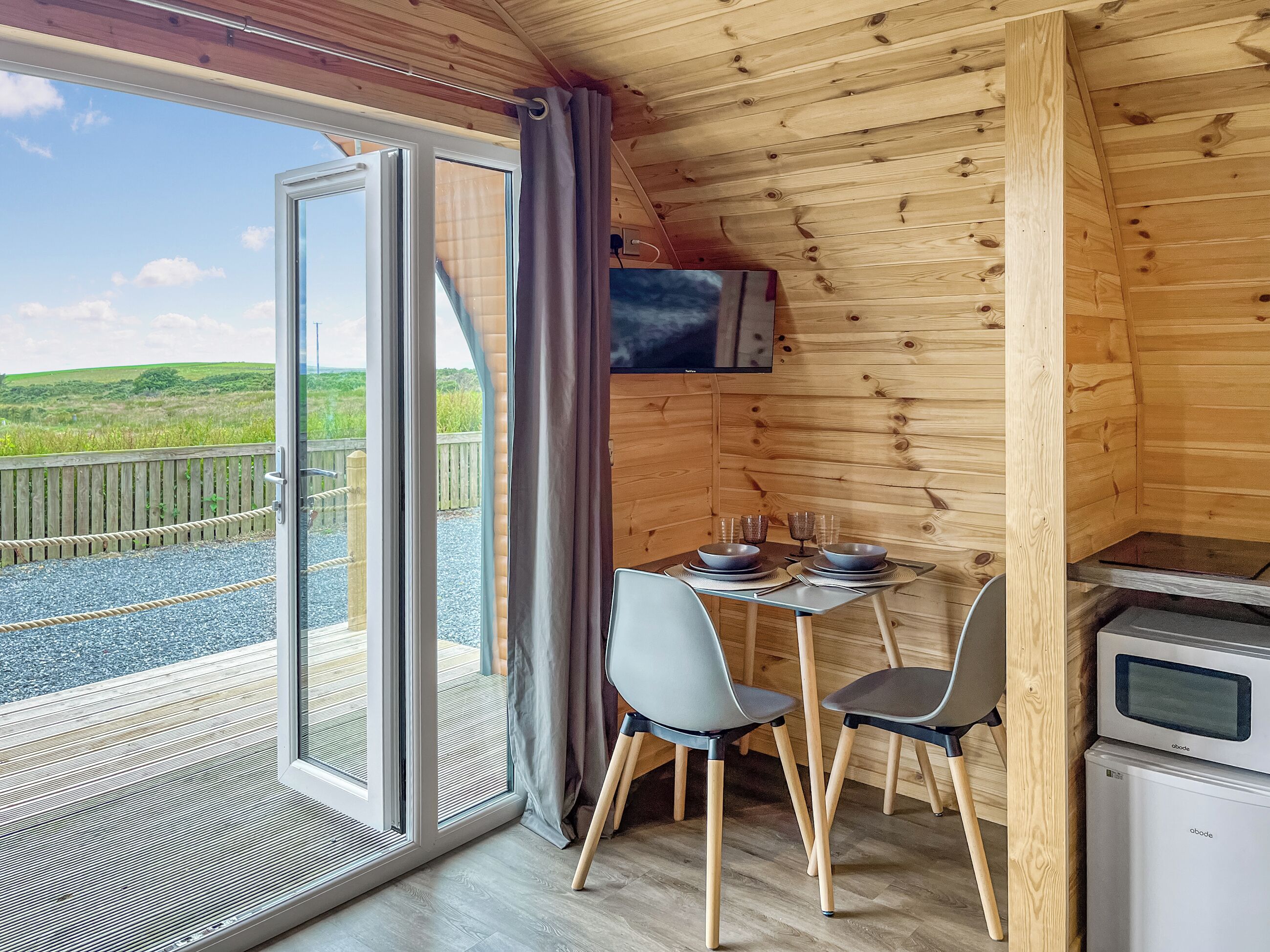 A wooden cabin with a dining area, a TV mounted on the wall, and a view of the outdoors.