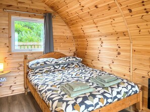 A wooden cabin room with a bed, a window with curtains, and a view of greenery outside.