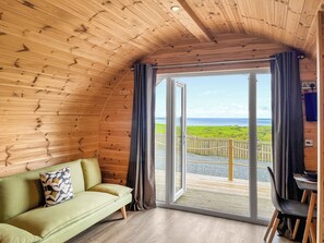 Ein gemütliches Blockhaus-Interieur mit Holzbalkendecke, einem grünen Sofa mit gemustertem Kissen und einem Tisch mit Stühlen. Eine große Schiebeglas-Tür führt auf eine Terrasse im Freien mit Blick auf das Meer und die grüne Landschaft.