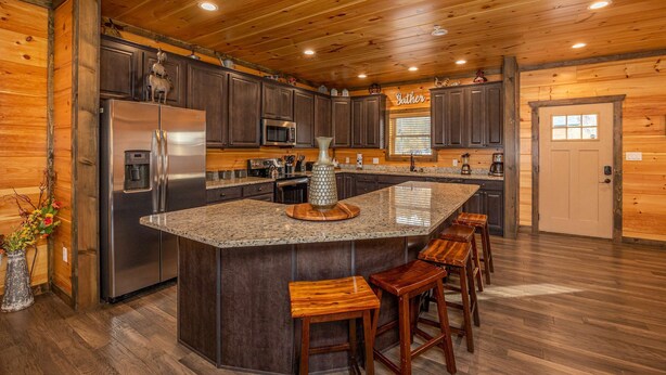 A kitchen with wooden cabinets, a stainless steel refrigerator, a granite countertop, and a bar stool area.