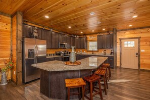 A kitchen with wooden cabinets, a stainless steel refrigerator, a granite countertop, and a bar stool area.