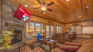 A cozy cabin interior with a fireplace, wooden walls, a ceiling fan, and a mounted TV displaying channel options.