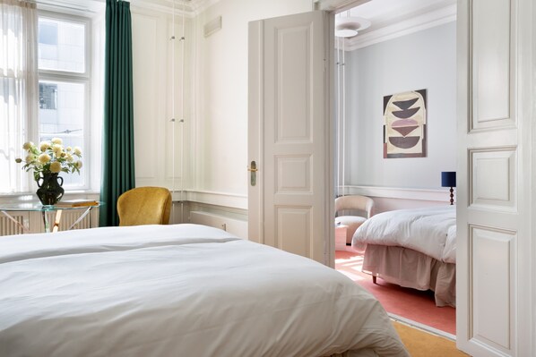 Habitación de hotel con dos camas, una zona de estar con una silla, una mesa con un jarrón de flores y vistas a la ciudad a través de una ventana con cortinas verdes.
