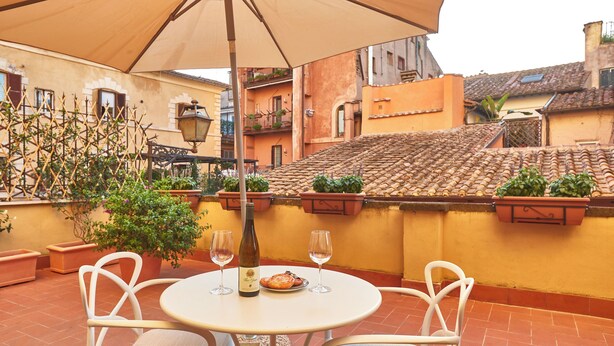 Mesa redonda con dos sillas, una botella de vino y un plato de comida bajo un paraguas grande en una terraza con baldosas de terracota y plantas en macetas.