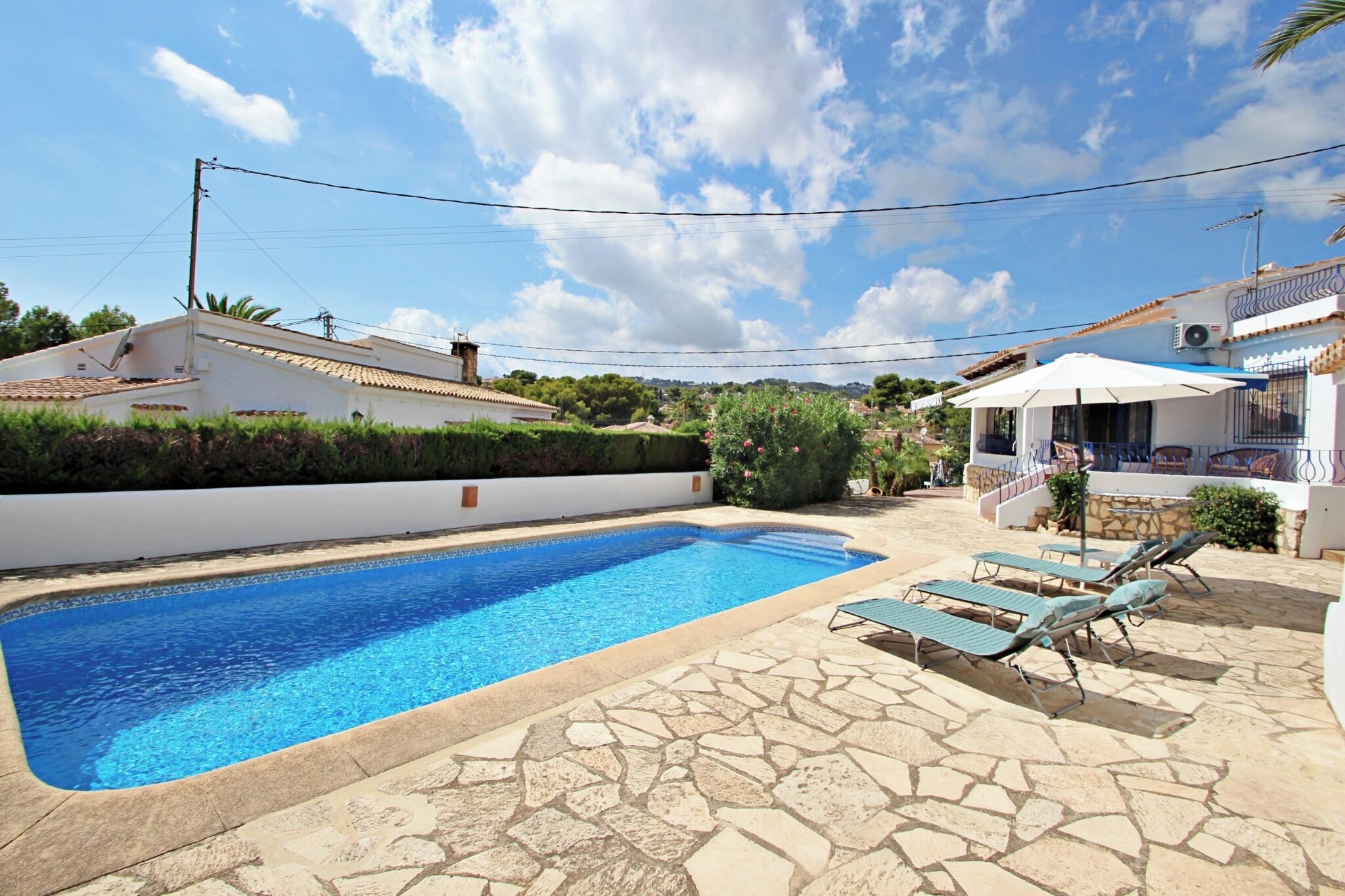 Une piscine entourée de chaises longues, une terrasse en pierre et un bâtiment blanc surmonté d’un toit bleu.