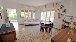 A room with a bed, a dining table with chairs, a small kitchen area, and a view of the outdoors.