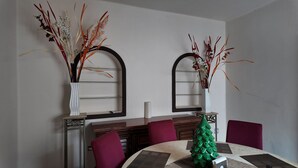 A dining area with two white vases with dried plants, a green Christmas tree, and purple chairs around a round table.