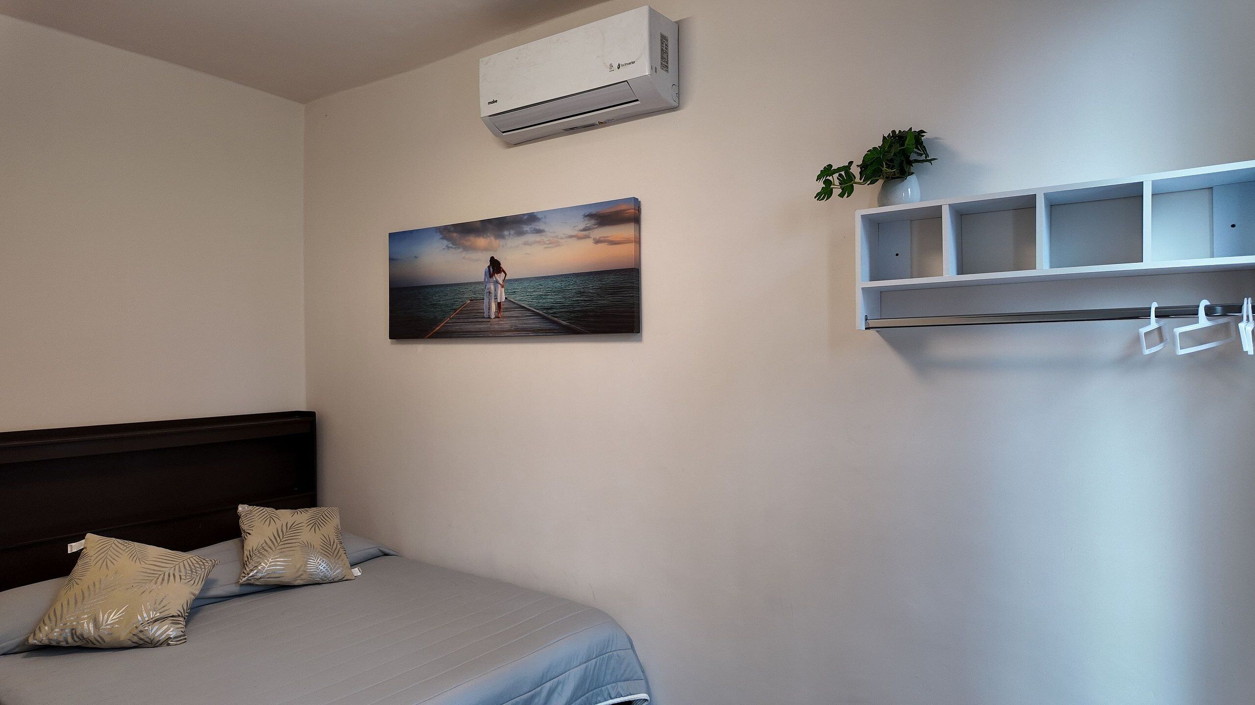 Un dormitorio con una cama, aire acondicionado de pared, un cuadro con una vista de atardecer sobre el agua y una pequeña estantería con una planta.