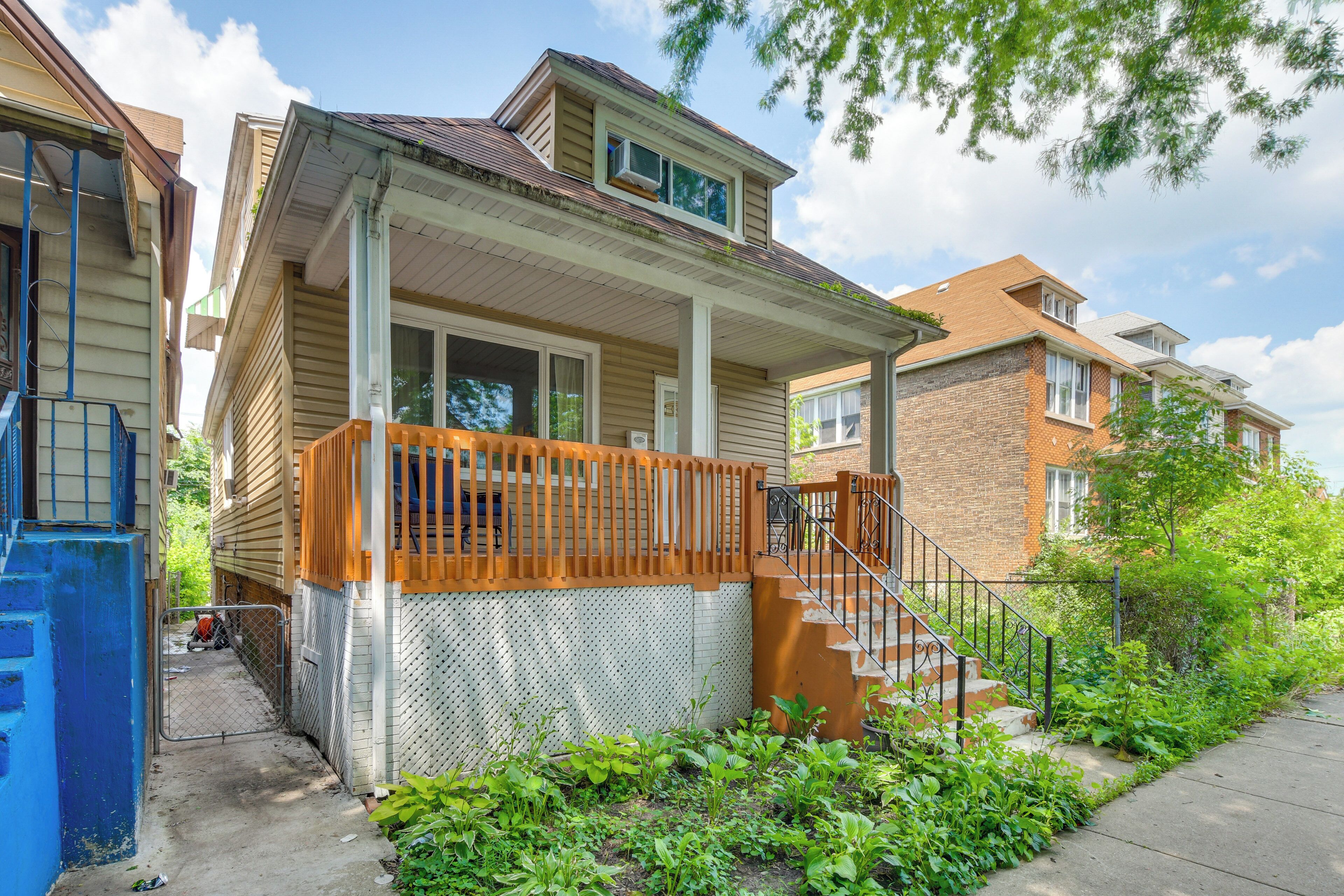A two-story residential building with a covered porch, a brick facade, and a small front yard with plants.