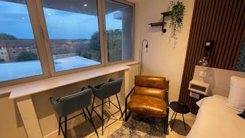 Un dormitorio con un ventanal grande, un escritorio con dos sillas, un sillón de cuero marrón, una mesita de noche y una cama.