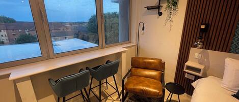 Une chambre avec une grande fenêtre, un bureau avec deux chaises, un fauteuil en cuir marron, une table de chevet et un lit.