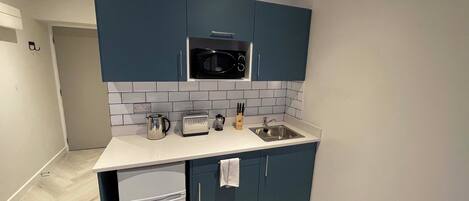 A compact kitchen with dark blue cabinets, a white countertop, a microwave, a toaster, a kettle, and a sink.