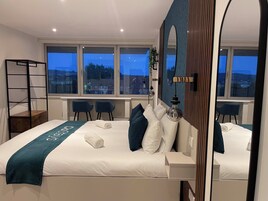 A modern hotel room with a large bed, two chairs, a desk, and a view of the city at dusk.