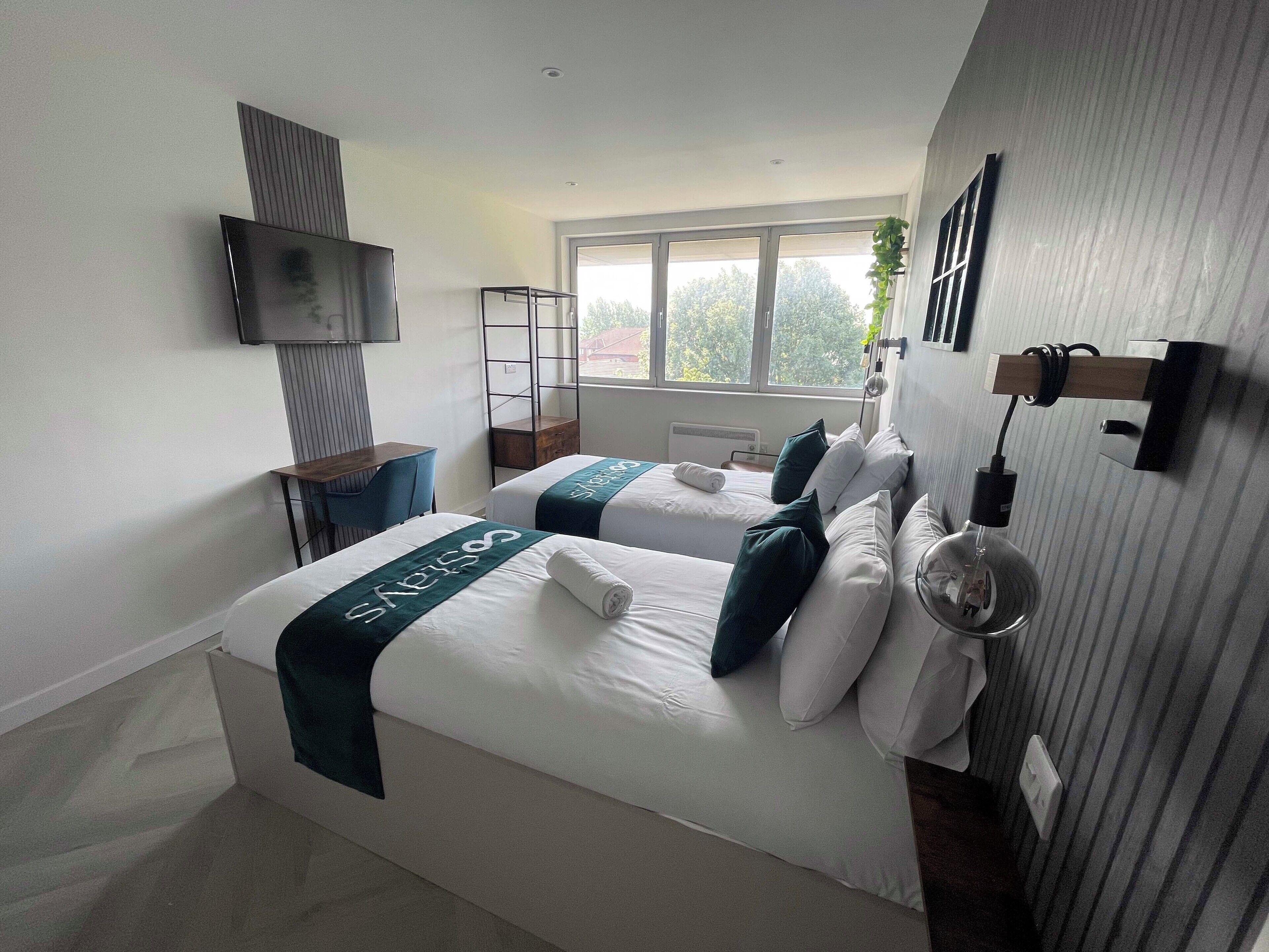 A modern hotel room with two beds, a TV mounted on the wall, a desk, and a large window offering a view of the outdoors.